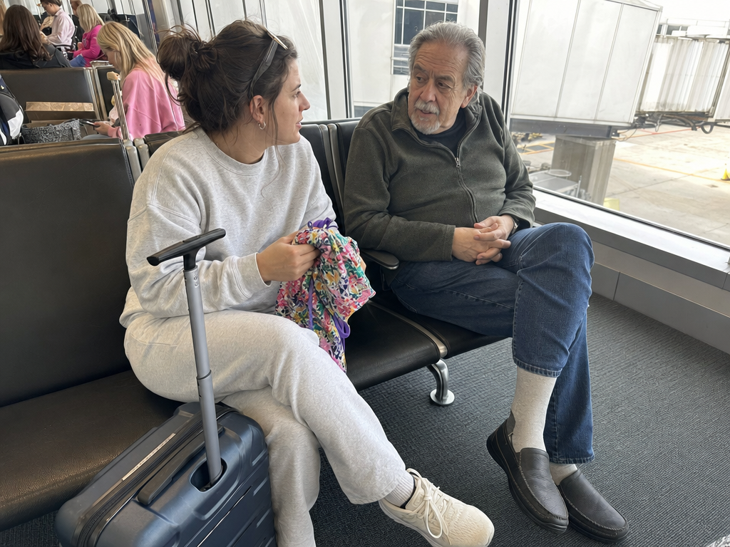 A woman holds a swimsuit beside her suitcase while speaking with her father-in-law at an airport gate.